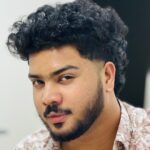 Close-up portrait of a young man with curly hair and a beard, looking confidently at the camera.